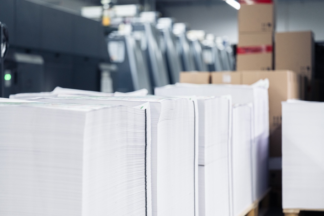 Große Papierstapel mit sauber geschnittenen Kanten sind auf Paletten in der Druckerei Esslinger Druck in Villingen-Schwenningen platziert. Die Druckbögen sind exakt übereinandergelegt und bereit für die Weiterverarbeitung oder Verpackung. Hinter den Stapeln stehen mehrere Druckmaschinen in Reihe, was auf eine Serienfertigung hindeutet. Die Szene zeigt professionelle Papierverarbeitung im industriellen Umfeld.