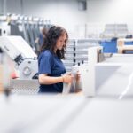 Eine Mitarbeiterin der Druckerei Esslinger Druck in Villingen-Schwenningen steht an einer industriellen Weiterverarbeitungsmaschine und hält einen Stapel frisch produzierter Mappen in den Händen. Sie trägt ein blaues T-Shirt und prüft konzentriert die ausgerichteten Druckprodukte vor der Weiterverarbeitung. Im Hintergrund sind weitere Druckmaschinen, Stapelboxen und Produktionsanlagen zu erkennen. Das Bild zeigt einen professionellen Arbeitsschritt in der Druckproduktion innerhalb einer hellen Fertigungshalle.