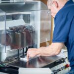 Ein älterer Mann mit Brille bedient eine industrielle Papierbohrmaschine in der Druckerei Esslinger Druck in Villingen-Schwenningen. Er richtet einen Stapel weißer Papierbögen präzise unter den Bohrköpfen aus, während die Maschine durch eine transparente Schutzabdeckung gesichert ist. Auf dem Arbeitstisch liegen weitere Papierstapel bereit, und Bedienelemente mit farbigen Knöpfen sind sichtbar. Die Szene zeigt einen professionellen Arbeitsschritt in der Weiterverarbeitung von Druckprodukten.